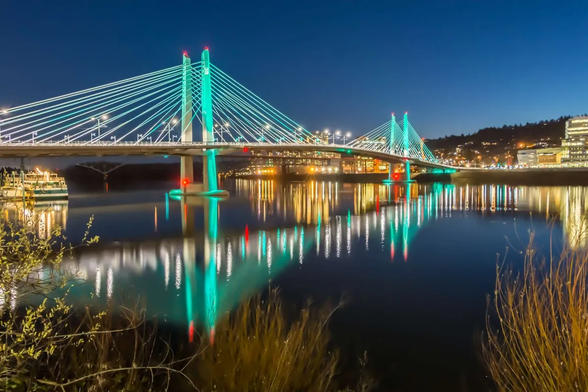 A glowing, illuminated bridge stretches across the water during evening hours, with city lights and reflections shimmering in the background. The lively atmosphere hints at bustling waterfront dining and nightlife options, along with opportunities for relaxing evening strolls.