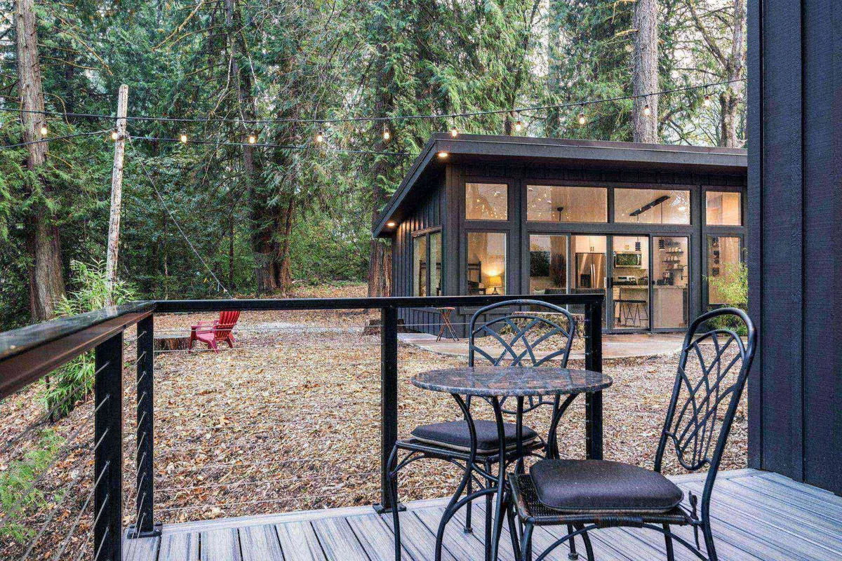 A tranquil evening setting with a modern cabin, cozy outdoor seating area, glowing string lights, and surrounding trees. The peaceful mood suggests both opportunities for private relaxation and small group socializing.