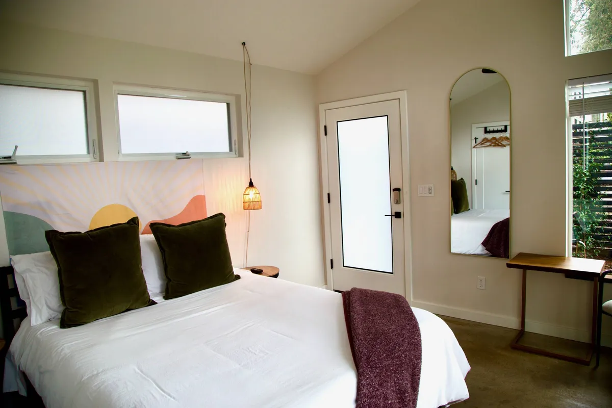 A serene bedroom scene showcasing a bed with decorative pillows, a pendant light feature, and a mirrored wall accent. Gentle morning light bathes the space, emphasizing a sense of calm and retreat.