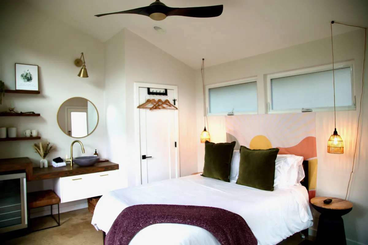 A tranquil bedroom with a plush bed adorned with decorative pillows, warm wood accents, hanging pendant lights, and a stylish wooden desk with a mirror. The space is filled with soft natural light, creating an inviting and serene atmosphere for relaxation or inspiration.