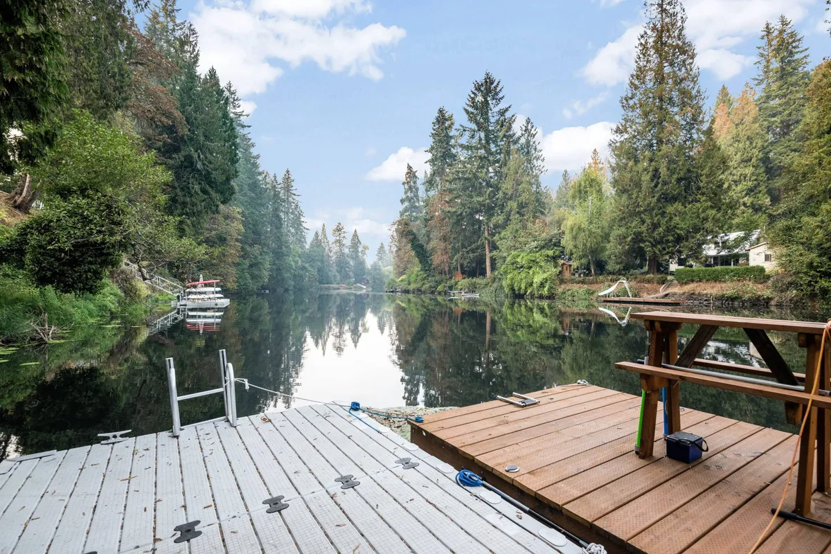 A calm river scene features a wooden dock stretching over still water, surrounded by lush trees. The dock is a focal point for relaxation or waterside activities, evoking a peaceful communal environment for both solitude and social engagement.