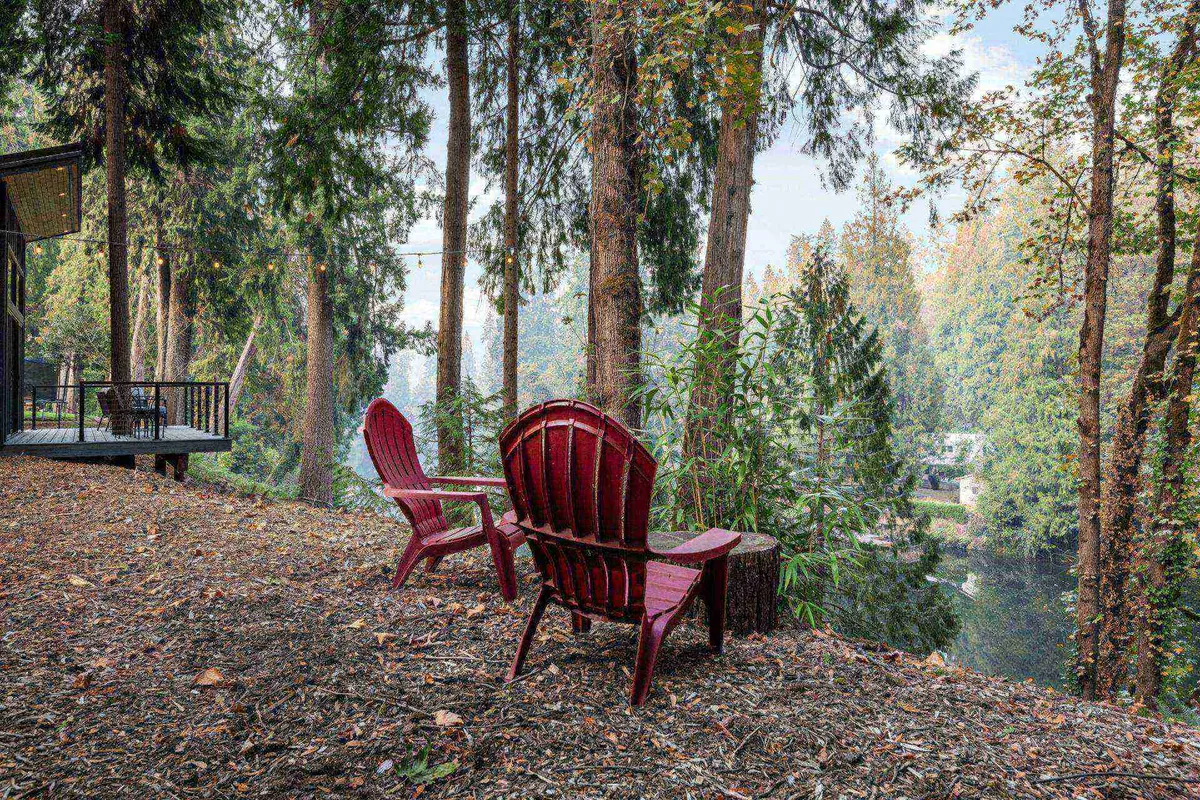 An exterior riverside setting with two classic red Adirondack chairs facing a calm river, framed by tall trees. The tranquil outdoor space is perfect for quiet moments, offering a direct connection to the natural setting described in the suite's features.