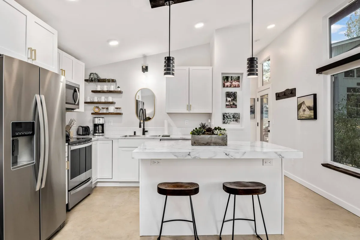 A bright kitchen area with marble countertops, stainless steel appliances, open shelving, and decorative plants. The scene feels contemporary and inviting, ideal for cooking, dining, and casual gathering.
