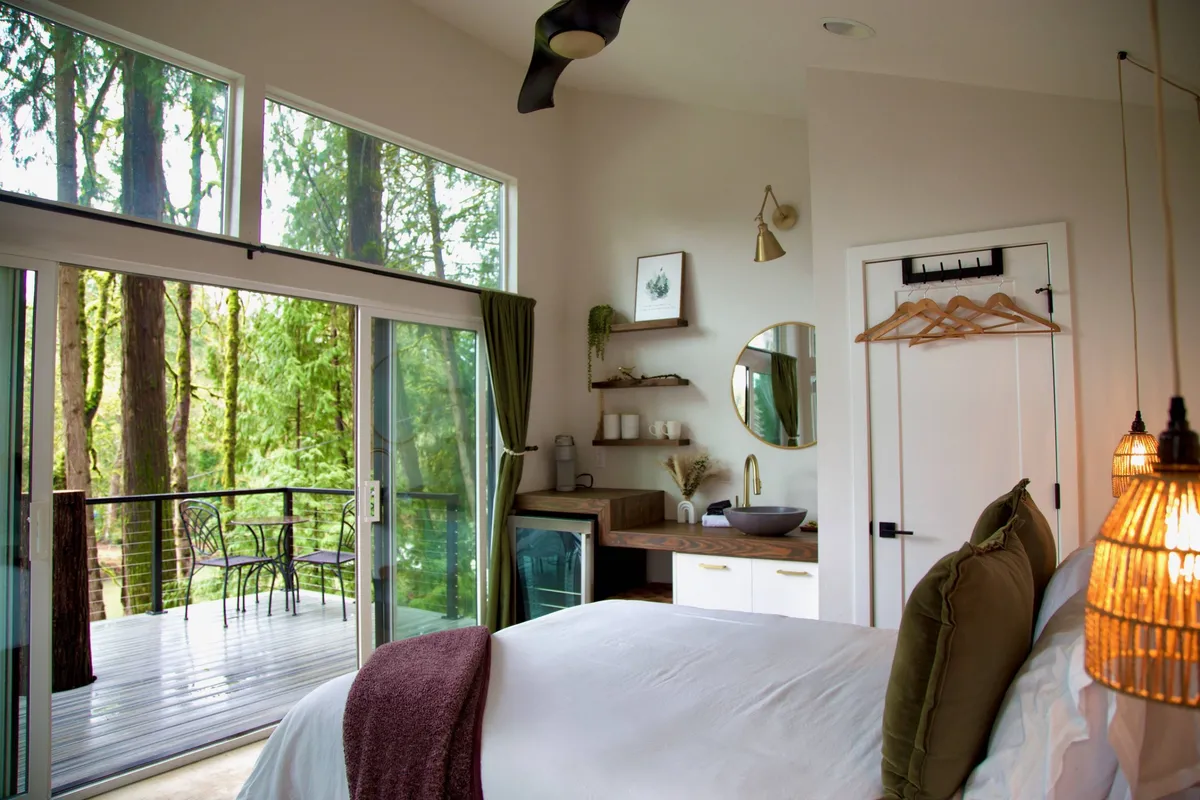 A tranquil bedroom scene with a plush bed, large windows overlooking lush trees, and stylish hanging light fixtures. The natural light fills the space, offering views of the surrounding forest and river, while the decor feels serene and restful.