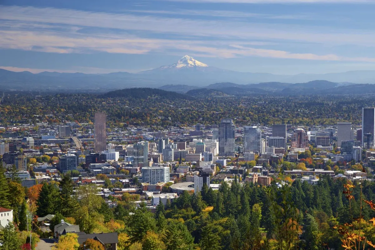 A sweeping view that captures Mount Hood in the distance, a vibrant city skyline, and colorful autumn foliage in the foreground. The scene transitions from urban structures to lush natural landscapes, hinting at the proximity of both outdoor adventures and city exploration.