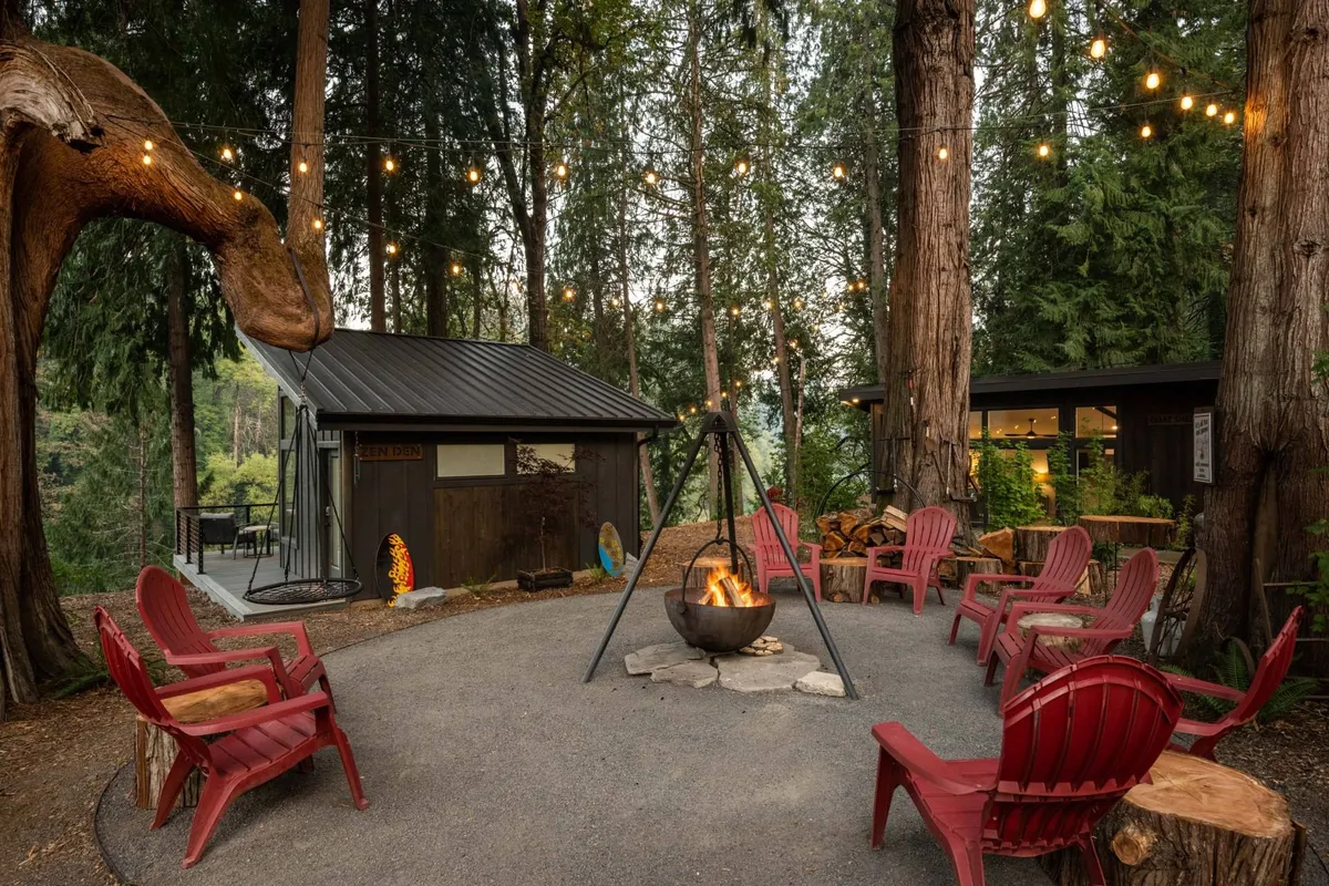 The image showcases the exterior of the Goat Shed cottage at dusk, featuring a glowing fire pit surrounded by bright red chairs, soft hanging string lights overhead, and a backdrop of lush, towering trees. The inviting scene suggests a perfect spot for evening gatherings, stargazing, and cozy relaxation in a woodland setting.