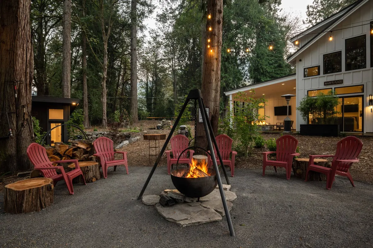 An inviting exterior scene featuring a fire pit surrounded by red Adirondack chairs, with a backdrop of a cabin-like structure and mature trees. The arrangement encourages communal gatherings and relaxing evenings outdoors.