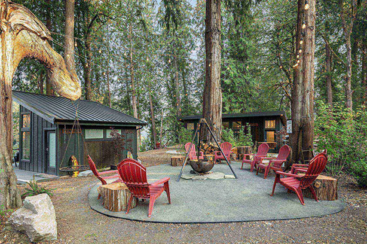 A cozy firepit area with adirondack chairs arranged around a glowing fire, framed by modern cabins and enveloped by tall trees. The communal atmosphere is evident as the space invites gatherings and storytelling under the stars.