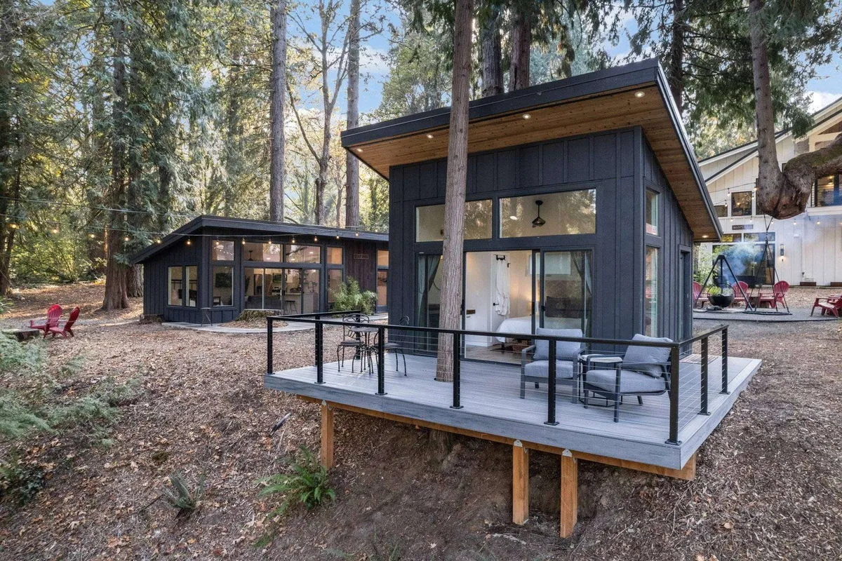 A modern cabin nestled among tall trees, with a patio seating area that overlooks the surrounding forest. The deck offers ample space to relax while taking in the tranquil natural setting.