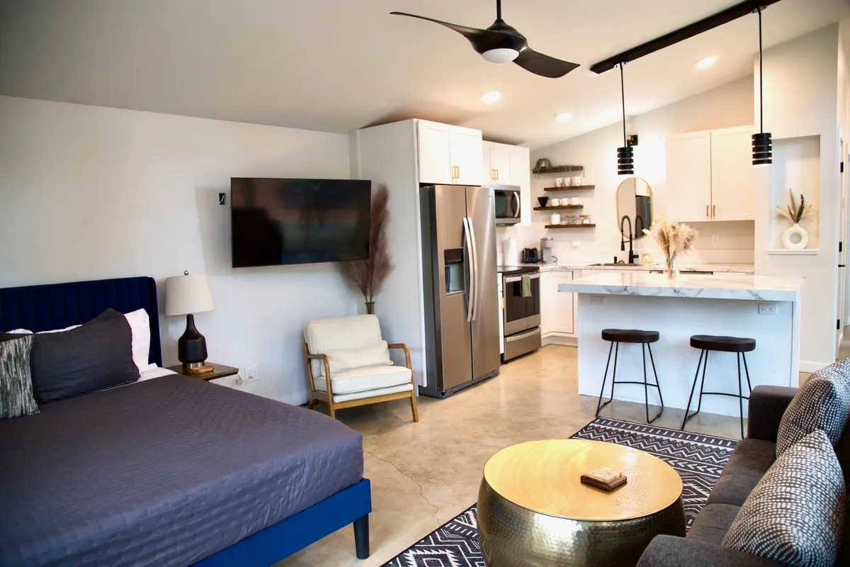 A spacious and stylish open-plan living area featuring a blue bed with decorative pillows, a golden round coffee table, modern kitchen appliances, stylish lighting fixtures, and a comfortable seating area. The living space is bright and welcoming, seamlessly blending the bedroom, kitchen, and relaxation zones.