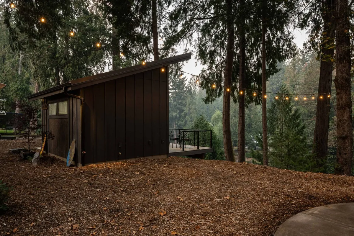 A modern cabin exterior is aglow with string lights, surrounded by mature trees and set against a tranquil natural backdrop. The outdoor gathering area exudes warmth and sociability, suggesting a space perfect for evening relaxation or group activities.