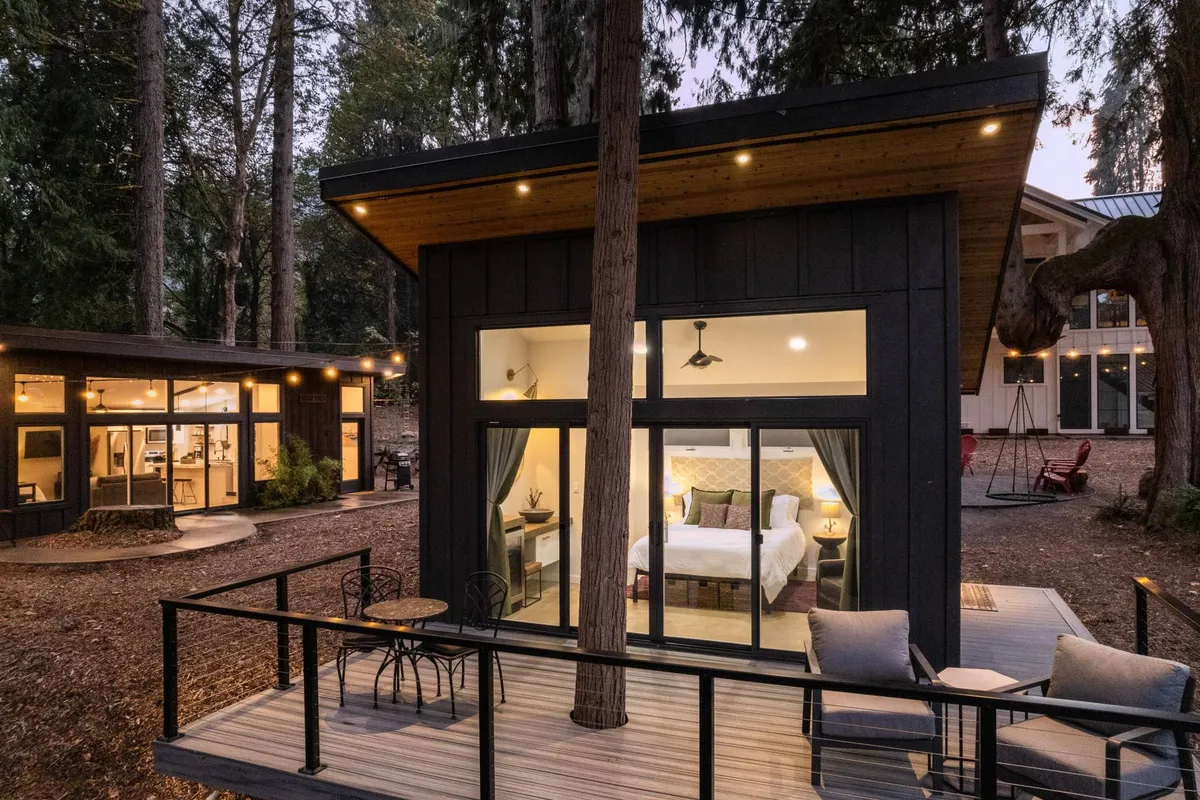 This image displays a tranquil deck area attached to a building, with comfortable outdoor seating set under the shade of mature trees. Large windows visually connect the indoor and outdoor spaces, while the natural surroundings evoke a sense of peace and connection with nature.