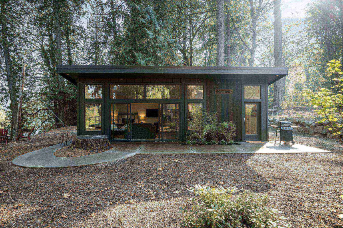 A modern cabin set amidst towering trees, featuring expansive glass windows that reveal a welcoming interior. The image also highlights a spacious outdoor patio equipped with a propane grill, surrounded by tranquil greenery.