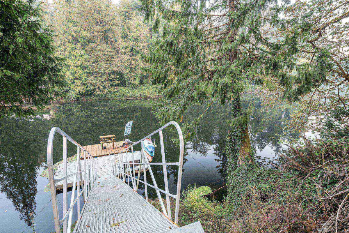 A metal dock extends over calm water, surrounded by tall trees and a wooden seating area, creating a perfect spot for nature observation or a peaceful stroll. The scene radiates tranquility and offers a direct connection to the water’s edge and forest environment.
